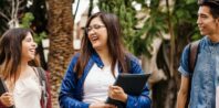 Three students are walking outdoors and smiling. They are Latino and Hispanic, holding notebooks and backpacks, appearing engaged in conversation. Tall trees and greenery are visible in the background, suggesting a campus or park setting.