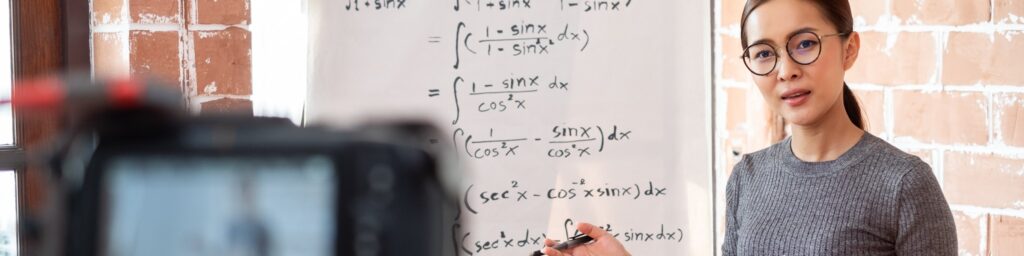 A person with glasses, wearing a gray shirt, stands near a whiteboard covered with calculus equations. Holding a marker and teaching strategies for understanding complex math concepts, they gaze towards the online audience through the camera. Brick walls are visible in the background.